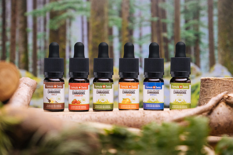 Seis aceites de CBD de espectro completo Formula Swiss con distintos aceites portadores y sabores, delante de un bosque