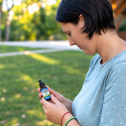 Mujer en un parque leyendo la etiqueta de un frasco de aceite de CBD, con camiseta azul de lunares y pulseras